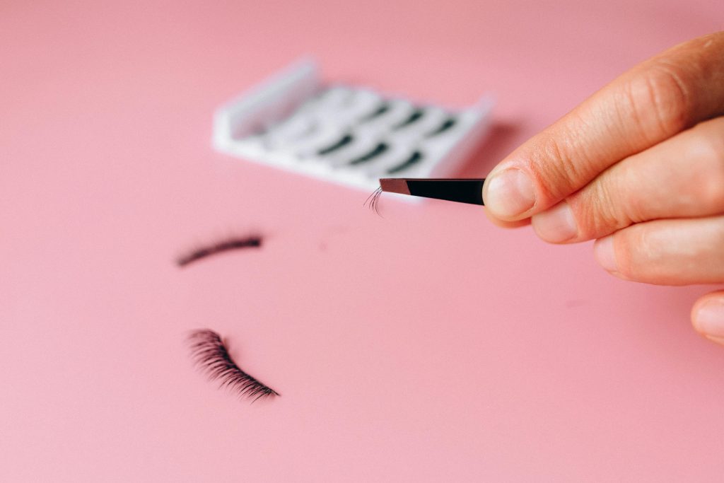 Close-up of applying false eyelashes with tweezers over a pink background.